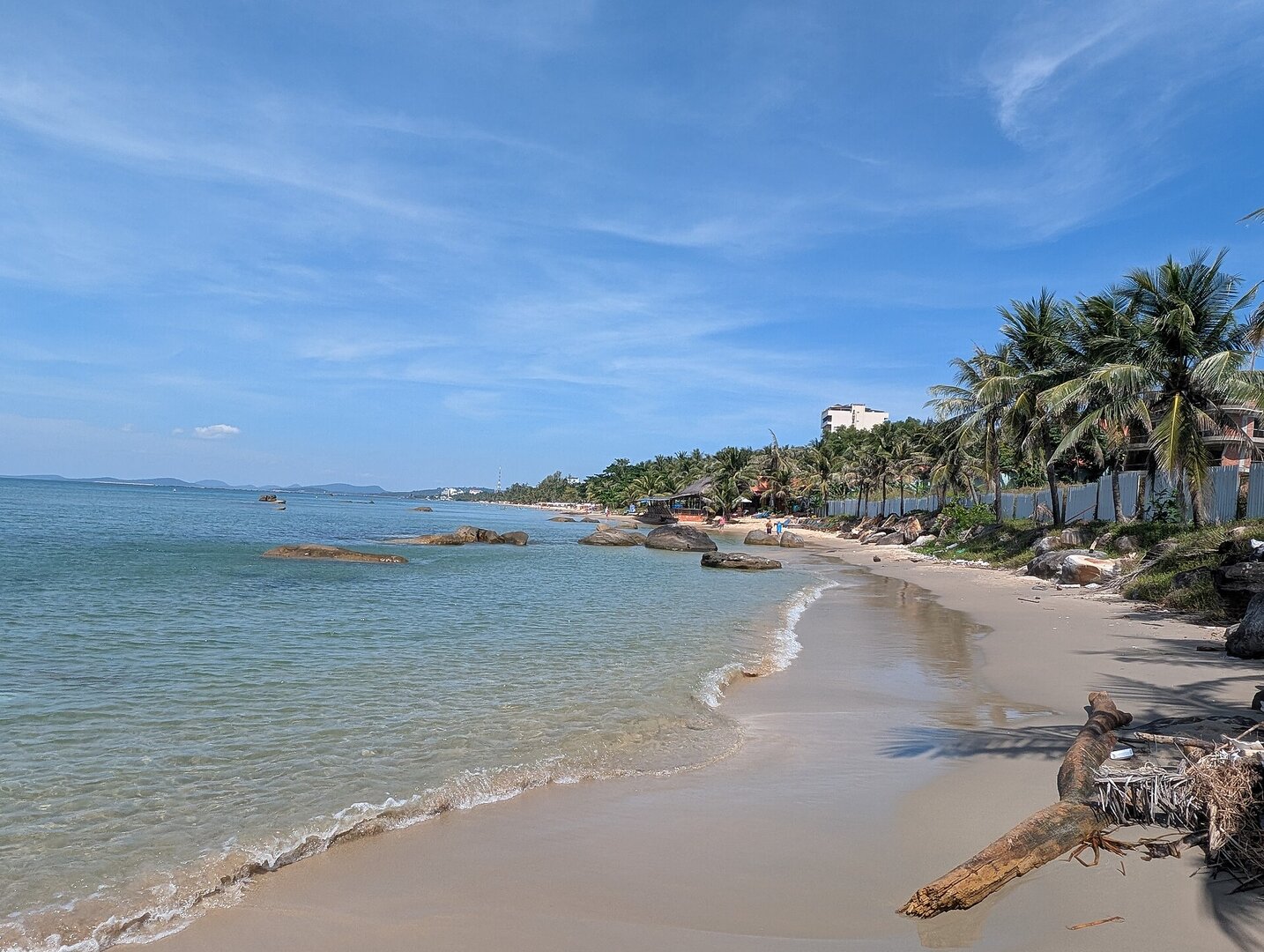 Bãi biển bờ tây Phú Quốc dưới trời xanh: nước trong xanh lặng sóng, những tảng đá tròn ven bờ và hàng dừa nghiêng ra biển.